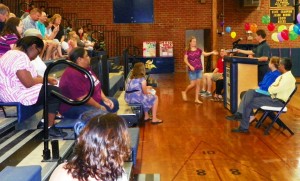 The second photo, shows a sixth-grade student approaching the podium to receive a certificate from her teacher, Stephen Jones, during the awards presentations for her grade level.